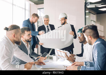 Focused indian, arabian and caucasian engineers, with one of them wearing hardhat, discussing construction graphs and blueprints in construction compa Stock Photo