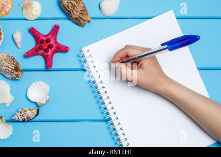 Blank notepad and hand with a pen different marine items on blue painted wooden background. Sea objects on wooden planks. Selective focus. Place for t Stock Photo
