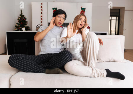 Cheerful shocked young couple sitting together on a couch at home, watching TV Stock Photo