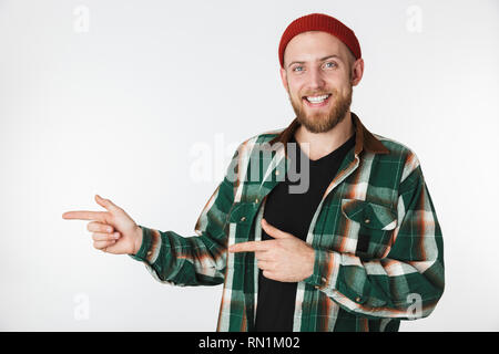 Portrait of young bearded guy wearing hat and plaid shirt smiling while pointing fingers aside isolated over white background Stock Photo