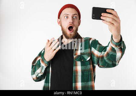 Image of joyful guy wearing hat and plaid shirt taking selfie photo on cell phone while standing isolated over white background Stock Photo