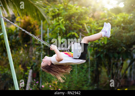 Child swinging on playground on sunny summer day in a park. Kids swing. School or kindergarten yard and play ground. Little girl flying high in the ai Stock Photo
