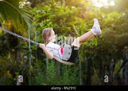 Child swinging on playground on sunny summer day in a park. Kids swing. School or kindergarten yard and play ground. Little girl flying high in the ai Stock Photo