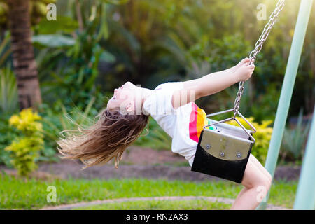 Child swinging on playground on sunny summer day in a park. Kids swing. School or kindergarten yard and play ground. Little girl flying high in the ai Stock Photo