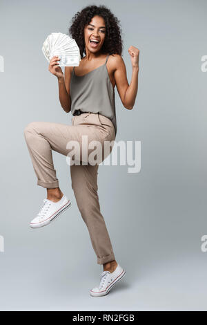 Happy young african woman standing isolated over gray background, holding bunch of money banknotes, celebrating Stock Photo