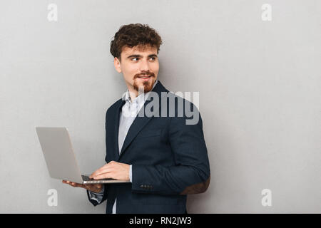 Handsome confident young businessman wearing a jacket standing isolated over gray background, working on laptop computer Stock Photo