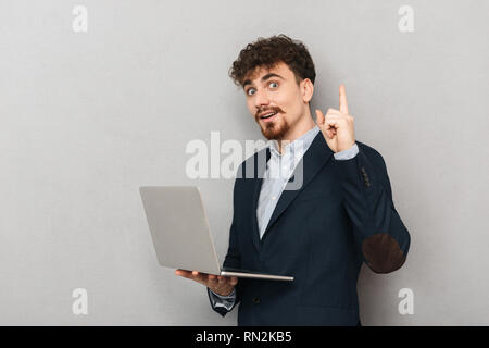 Pensive young businessman wearing suit standing isolated over gray background, working on laptop computer Stock Photo