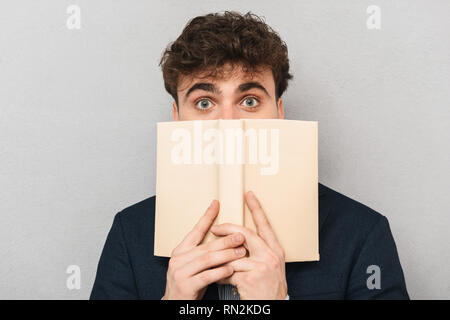 Surprised young businessman wearing suit standing isolated over gray background, covering his face with a book Stock Photo