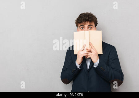 Surprised young businessman wearing suit standing isolated over gray background, covering his face with a book Stock Photo