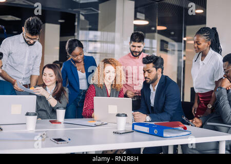 Creative multiracial business team discussing project, standing around workplace, while two co-workers with portable computers looking for needed info Stock Photo