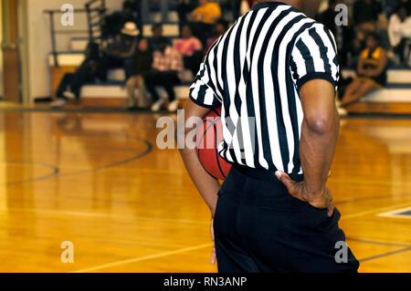 Referee stands mid court holding basketball under his arm.  He is wearing the usual black and white striped uniform. Stock Photo