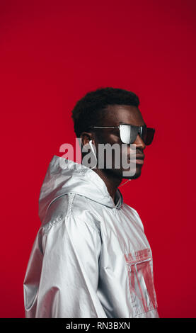 African man in futuristic outfit against red background. Man in stylish sunglasses,  silver hoodie and earphones. Stock Photo