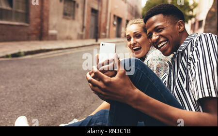 Man taking selfie with his female friend sitting outside by the street. Cheerful friends making a selfie outdoors. Stock Photo