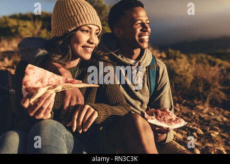 Young man and woman sitting on mountain trail eating pizza and looking at the view. Couple on hiking trip eating pizza and admiring the view. Stock Photo