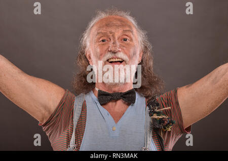 Old homeless but cheerful beggar with arms outstretched, wearing outworn jeans, black gloves and hat Stock Photo