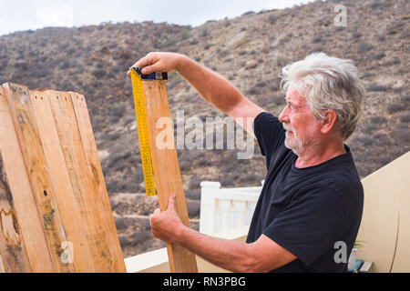 Adult retired man work outdoor like carpenter taking measure dimensions of wooden table - building and reparing or restoring in leisure activiy time - Stock Photo