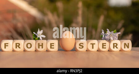 One Egg and dice form the German words 'FROHE OSTERN' ('HAPPY EASTER' in English) on a wooden table with flowers. Stock Photo