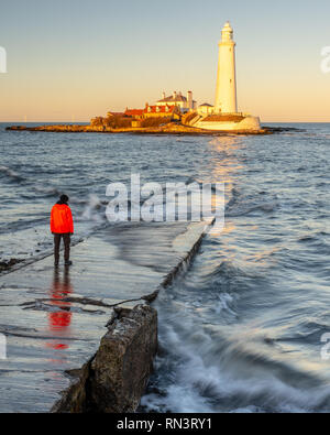 A man stands on the St Mary's Island Causeway while the setting sun illuminates St Mary's Lighthouse on the coast of Whitley Bay in Tyneside. Stock Photo