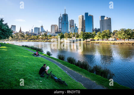 2nd January 2019, Melbourne Australia : Man reading a book on Yarra riverbank promenade grass in central Melbourne and Melbourne CBD skyline in Victor Stock Photo