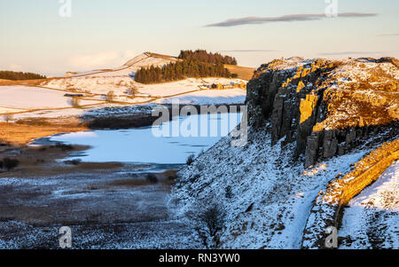 Winter snow lies on fields and crags beside the frozen lake of Crag Lough, while hikers walk along Hadrian's Wall path in Northumberland. Stock Photo