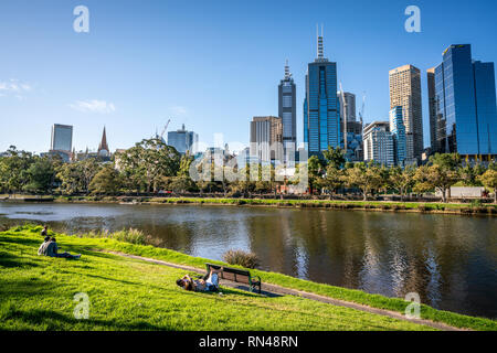 2nd January 2019, Melbourne Australia : People resting on Yarra riverbank promenade grass in central Melbourne and Melbourne CBD skyline in Victoria A Stock Photo
