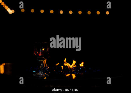 A glass of red wine sitting on the rim of fire pit with the flames reflecting thru the glass.  There is bokeh lights in the black background. Stock Photo