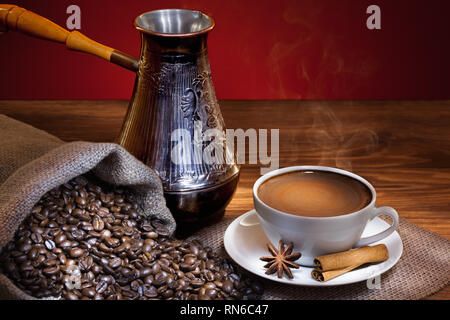 White cup of hot coffee with roasted beans and turkish coffee pot on wooden table. Stock Photo