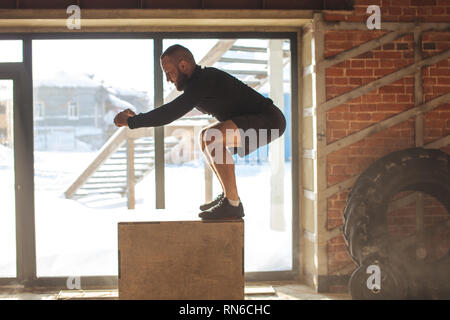 Box jump plyo exercise - young caucasian man doing functional workout at the well-lit gym in a sunny day, motion shot in touchdown phase. Stock Photo