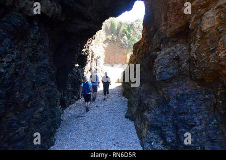 Three Hikers Walking through the Natural Arch in the Sandstone Cliff on Dixcart Bay on the Island of Sark, Channel Islands, UK. Stock Photo