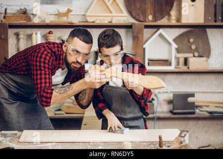 Father and little son testing hand-made wood sword evenness at workshop. Stock Photo