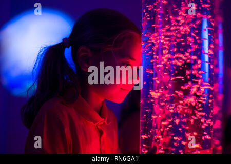 Child in therapy sensory stimulating room, snoezelen. Child interacting with colored lights bubble tube lamp during therapy session. Stock Photo