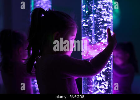 Child in therapy sensory stimulating room, snoezelen. Child interacting with colored lights bubble tube lamp during therapy session. Stock Photo
