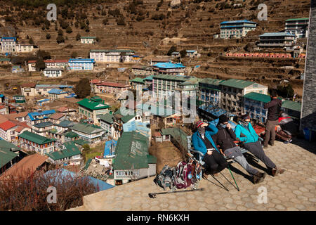 Nepal, Namche Bazaar, walkers wearing cold weather clothing resting on seat in sunshine with elevated view of town Stock Photo