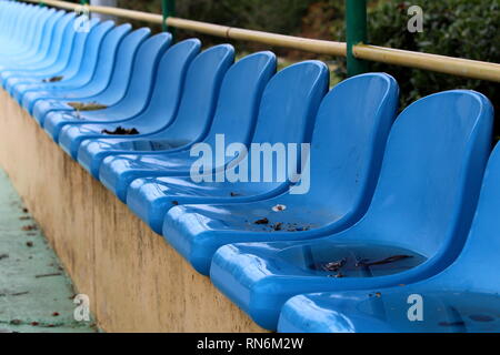 Row of blue plastic seats mounted on top of concrete bleachers with metal fence and trees in background next to basketball court on wet rainy day Stock Photo