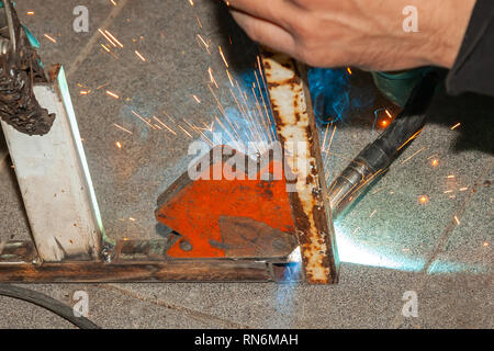 An experienced person performs work with a welding machine, fixing metal parts, removing blue smoke and yellow sparks and lightning in the industrial  Stock Photo