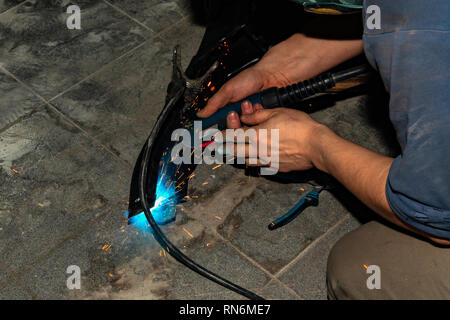 An experienced person performs work with a welding machine, fixing metal parts, removing blue smoke and yellow sparks and lightning in the industrial  Stock Photo