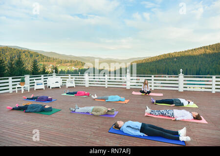 Yoga group practising relaxation on fresh air in mountains. People wearing in sportswear lying on special yoga mats, relaxing and enjoying nature. Stock Photo