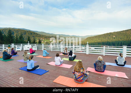Woman practicing yoga with coach, sitting in lotus pose on yoga mats. Instructor teaching yoga in mountains on fresh air. Yoga group relaxing and enjoying amazing view. Stock Photo