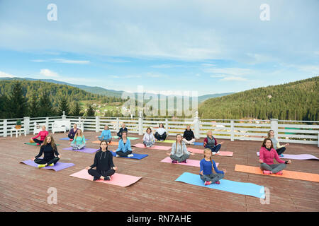 People relxing on yoga class, practicing meditation, sitting on mats. Group of women and girls meditating on fresh air in mountains. Women in sportswear enjoying amazing view. Stock Photo