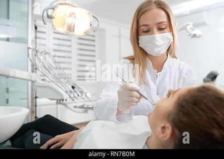 Professional dentist wearing medical mask, gloves and white uniform. Doctor treating teeth of patient, preventing caries. Woman lying in dentist chair. Stomatology concept. Stock Photo