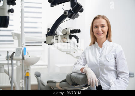 Charming woman working as stomatologist, posing at workplace. Beautiful female dentist sitting, leaning on dentist chair. Stomatologist in white uniform smiling, looking at camera. Stock Photo