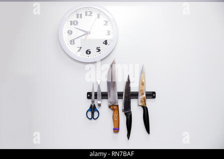 Round clock and knives with wooden and plastic handle and scissors on a magnetic holder on a white wall. Concept kitchen restaurant Stock Photo