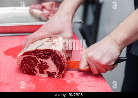 Closeup cook's hand cuts ribeye marbled beef steak with sharp knife on red plastic cutting board on metal table in restaurant kitchen. Concept steakho Stock Photo