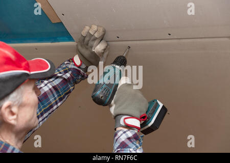 Drywall construction, attic renovation. Man fixing drywall suspended ceiling to metal frame using electrical screwdriver Stock Photo