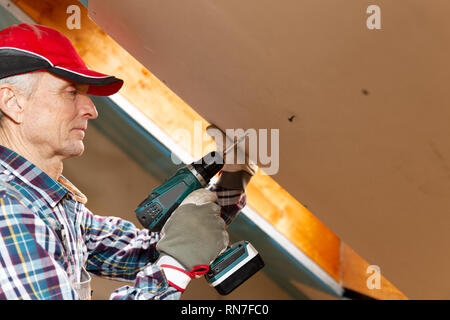 Drywall construction, attic renovation. Man fixing drywall suspended ceiling to metal frame using electrical screwdriver Stock Photo