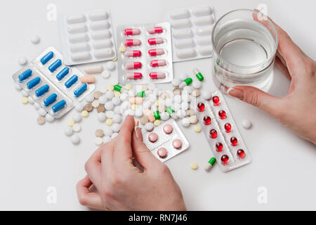 Heap of medication and pills with a glass of water and a hand holding a pill on white background. Top view. Stock Photo