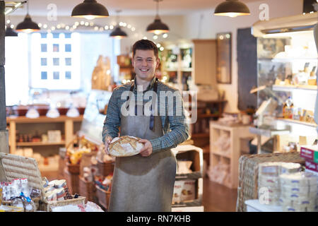 Portrait Of Smiling Male Owner Of Delicatessen Shop Wearing Apron Holding Loaf Of Bread Stock Photo