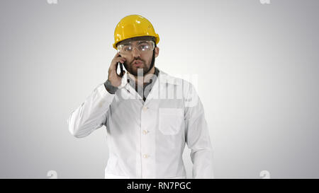 Engineer in protective glasses and hat walking, taking on the phone on gradient background. Stock Photo