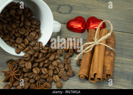 Roasted coffee beans spilling from a cup, two heart shaped candies, cinnamon with star anise spice on a rustic wooden table in a close up view Stock Photo