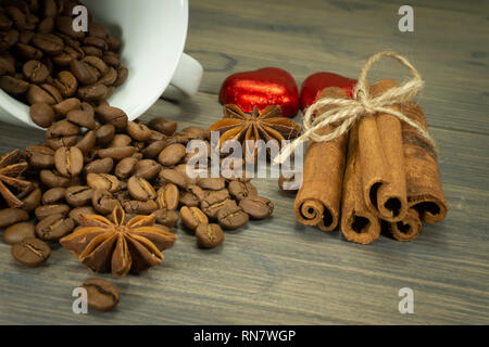 Roasted coffee beans spilling from a cup, two heart shaped candies, cinnamon with star anise spice on a rustic wooden table in a close up view Stock Photo